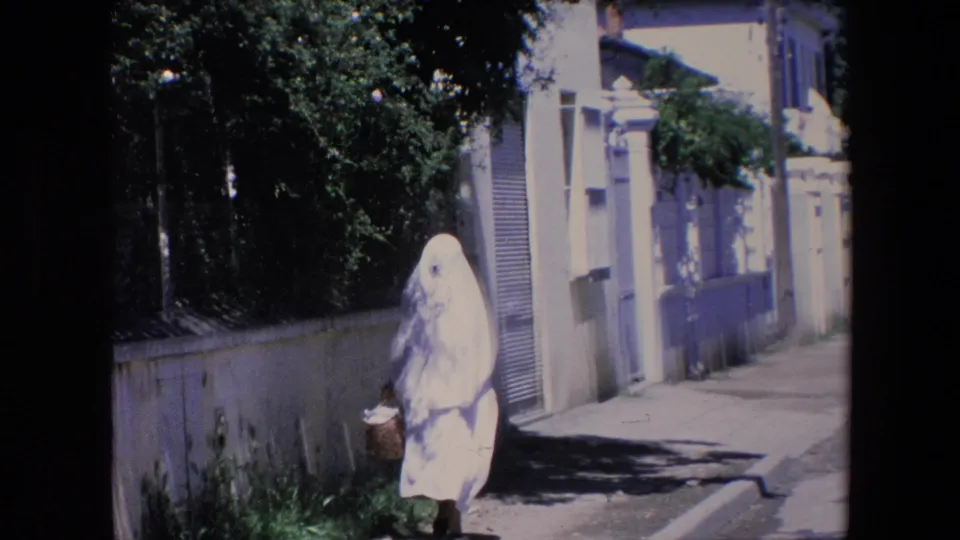 The woman in white passes nearer to the camera with a basket, framed by a low wall, tree shade, and pale residential buildings in the 1971 Algeria home-movie footage.