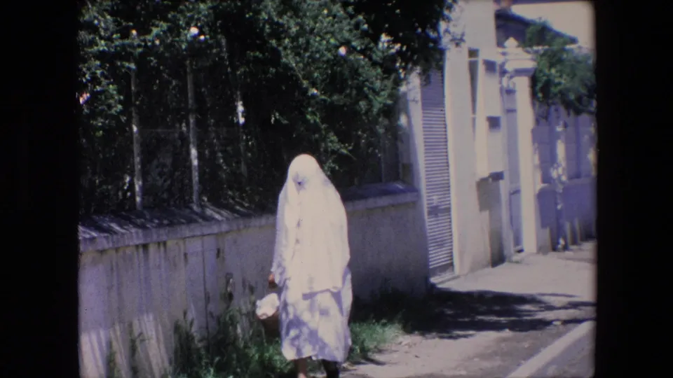 Near the end of the clip, the woman in a white full-body covering is close to the camera on the residential street, with bright stucco walls and tree shade visible behind her.