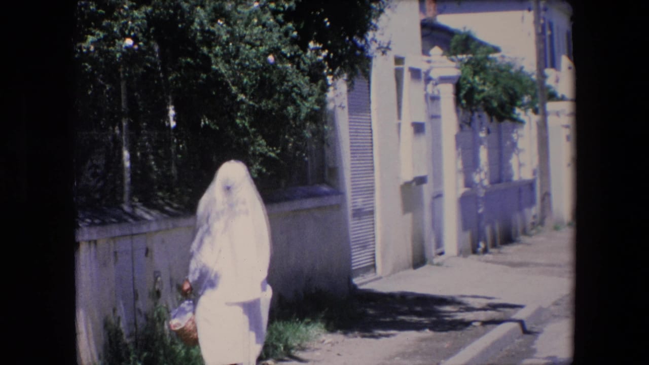 In Algeria in 1971 according to clip metadata, a woman in a white covering walks beside a wall and trees while carrying a woven basket on a bright street.