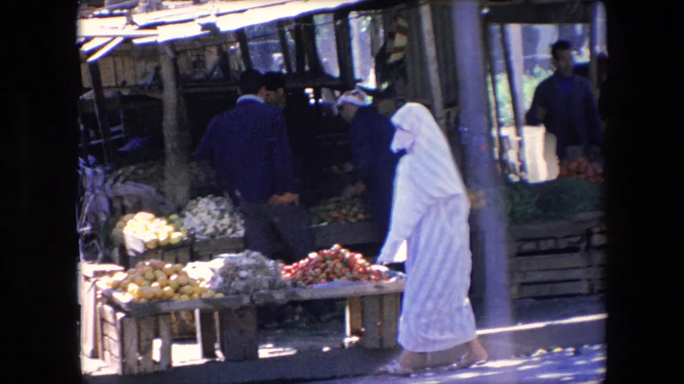 A market table piled with citrus, pale vegetables, and red produce sits in the foreground as a woman in white and several men gather near the stalls.