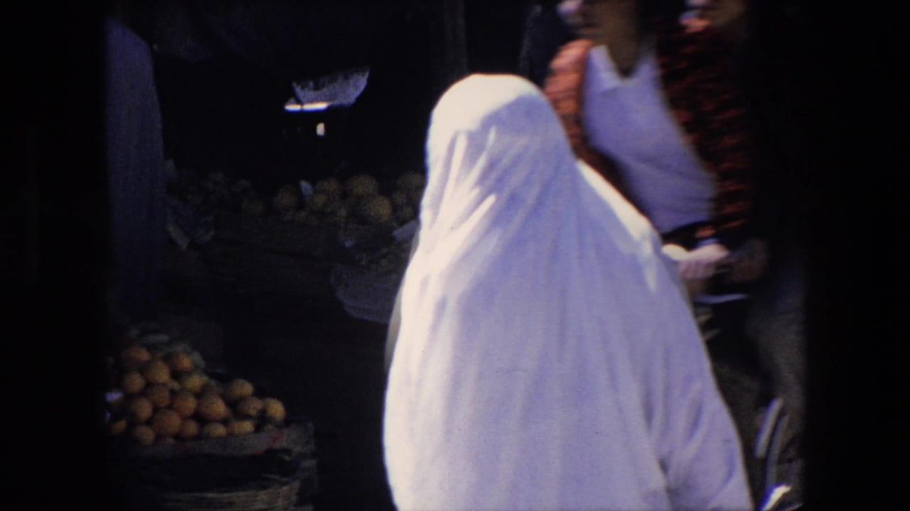 In metadata-supported 1971 Algeria footage, a woman in a white full-body covering walks near stacked produce while pedestrians and shadowed market stalls fill the background.