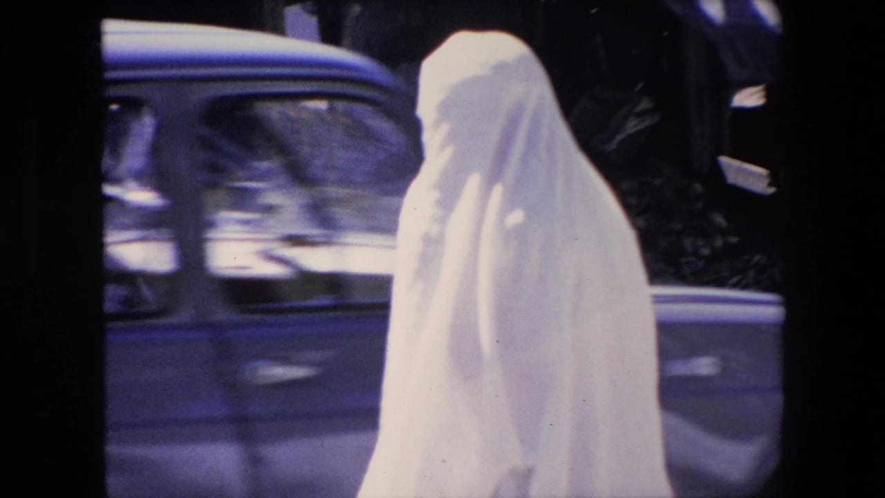 A woman in a white full-body covering walks along an Algerian urban street in 1971 as a car passes behind her and pedestrians remain partly visible near the frame edge.