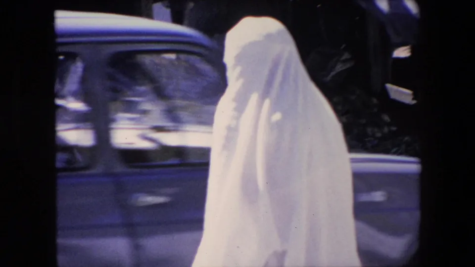 A woman in a white full-body covering walks along an Algerian urban street in 1971 as a car passes behind her and pedestrians remain partly visible near the frame edge.