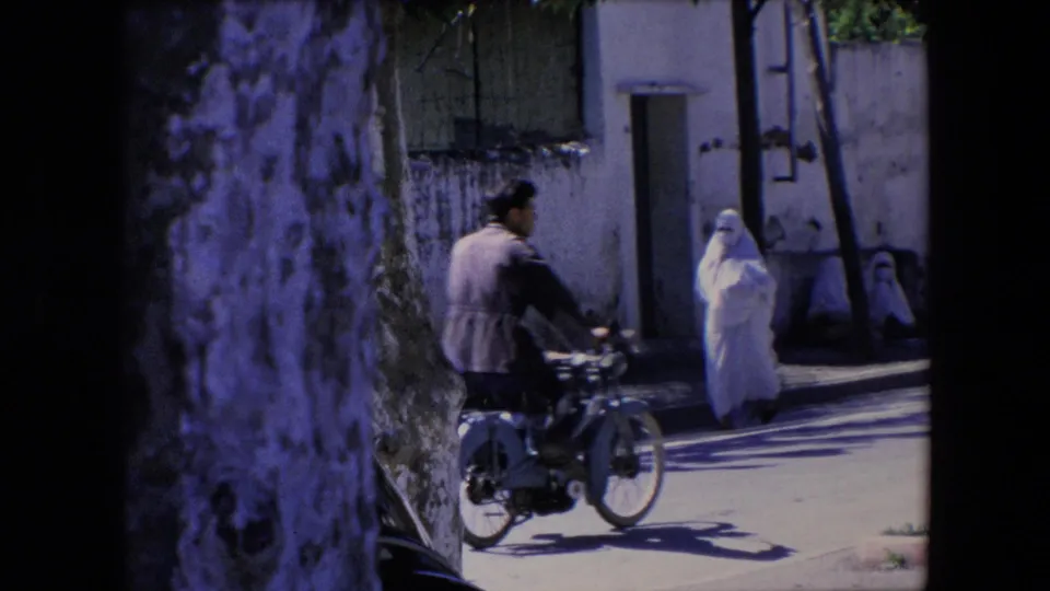 A motorbike rider moves through the foreground while a woman in a white full-body covering walks near a wall and doorway in the 1971 Algeria street scene.