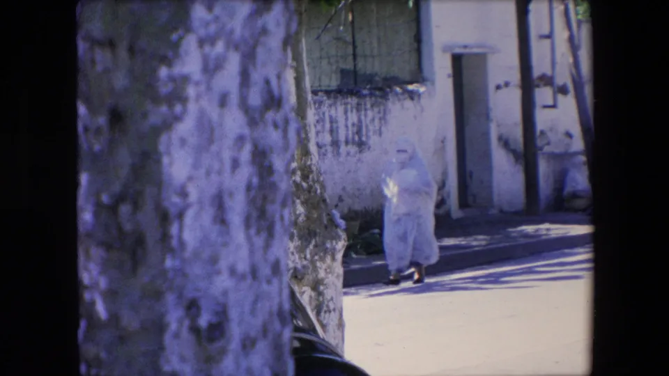 The woman in a white full-body covering continues along a sunlit sidewalk, partly framed by a tree trunk and exterior wall in metadata-supported 1971 Algeria footage.