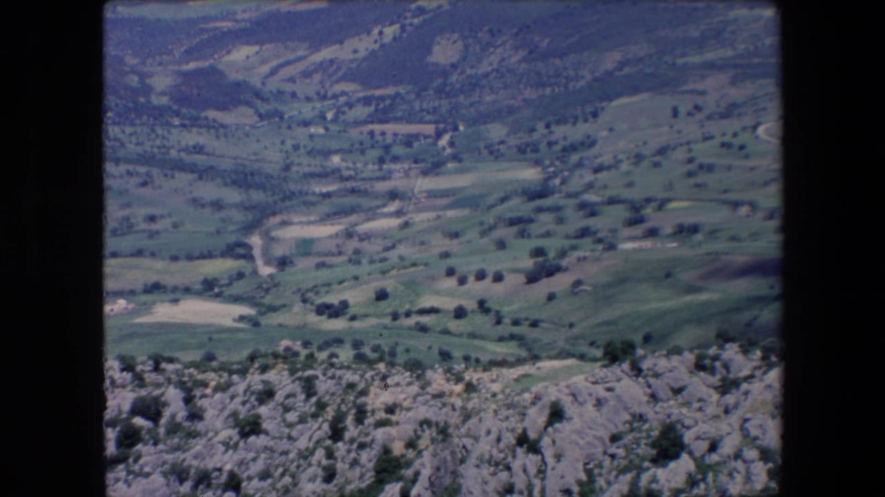 1971 Algeria 8mm frame showing a high overlook across rocky foreground terrain toward green hills, scattered trees, pale winding tracks, and distant rural structures.