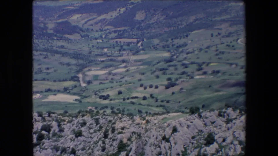 1971 Algeria 8mm frame showing a high overlook across rocky foreground terrain toward green hills, scattered trees, pale winding tracks, and distant rural structures.