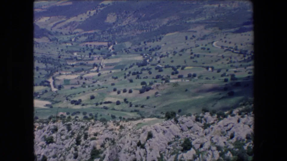 1971 Algeria 8mm frame with a distant rural valley, scattered trees, small structures, pale roads, and rocky terrain at the bottom edge of the view.