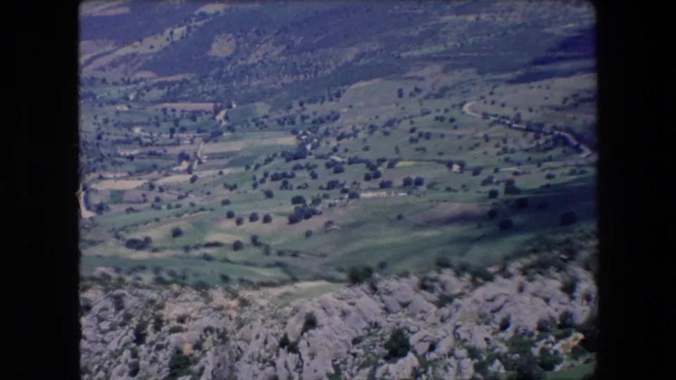 1971 Algeria 8mm frame looking across a mountainous rural valley with green fields, tree clusters, pale tracks, distant settlement forms, and rough rock in the foreground.