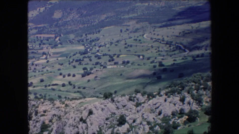 1971 Algeria 8mm frame showing a broad rural mountain valley from above, with rocky foreground, scattered trees, curving light-colored tracks, and layered hills beyond.