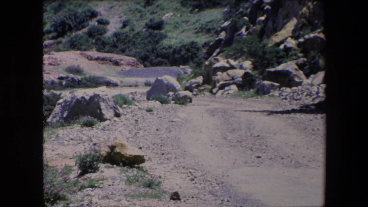 1971 Algeria 8mm frame with a pale dirt track running past large rocks, low shrubs, and rugged green slopes under bright daylight.