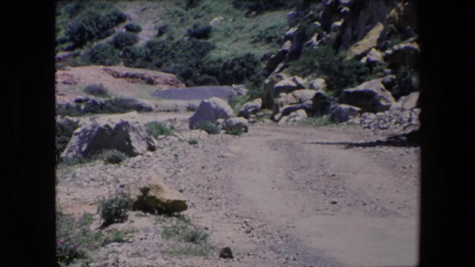 1971 Algeria 8mm frame with a pale dirt track running past large rocks, low shrubs, and rugged green slopes under bright daylight.
