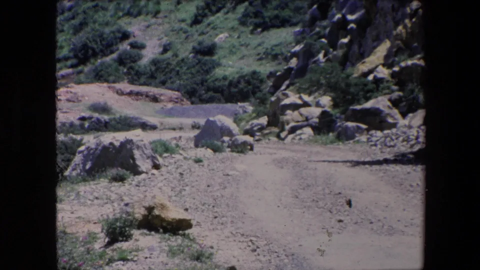 1971 Algeria 8mm frame showing the dirt path continuing through a rocky hillside with scattered boulders, scrub plants, and soft film grain.
