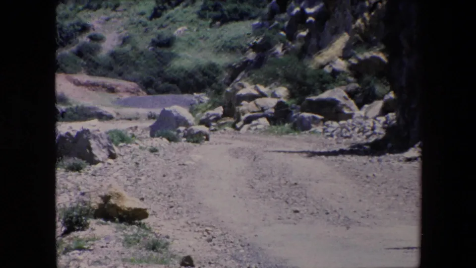 1971 Algeria 8mm frame of a quiet rural hillside where a dusty path passes between rocks, boulders, shrubs, and patches of green grass.