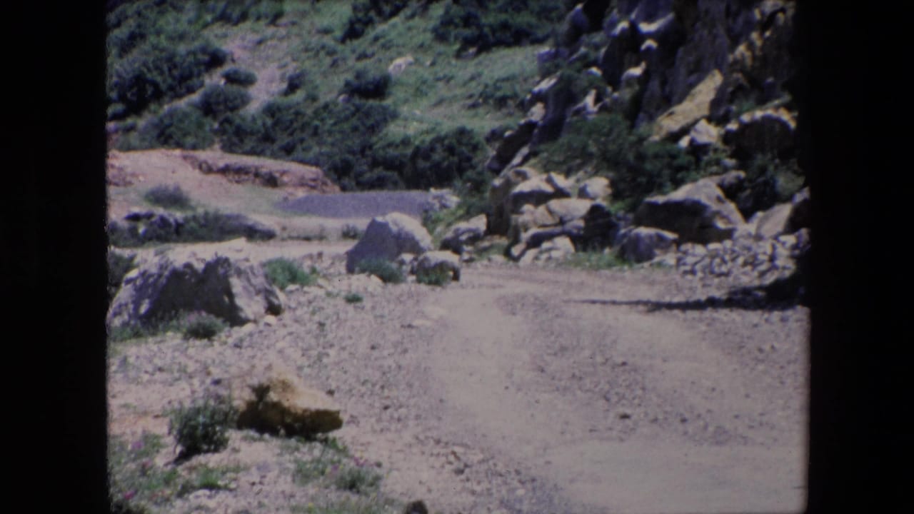 1971 Algeria 8mm frame showing a dusty rocky path curving past scrub vegetation and large boulders in a rugged hillside setting.