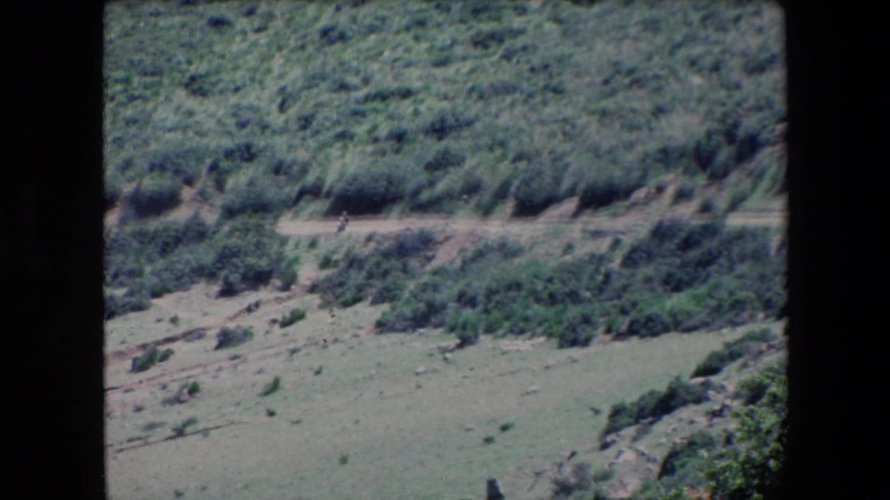 1971 Algeria 8mm frame looking across a shrub-covered hillside toward a pale dirt road with a very small distant person visible.