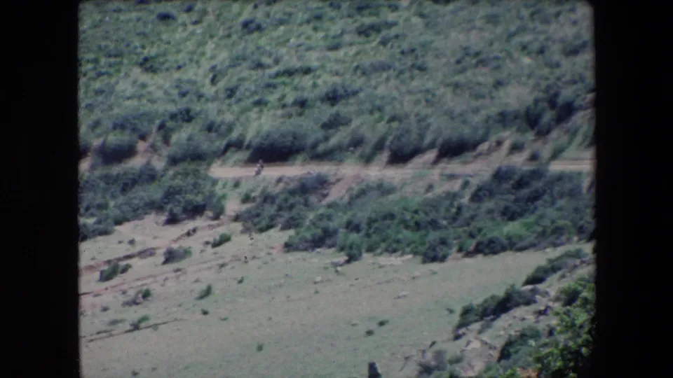 1971 Algeria 8mm frame looking across a shrub-covered hillside toward a pale dirt road with a very small distant person visible.