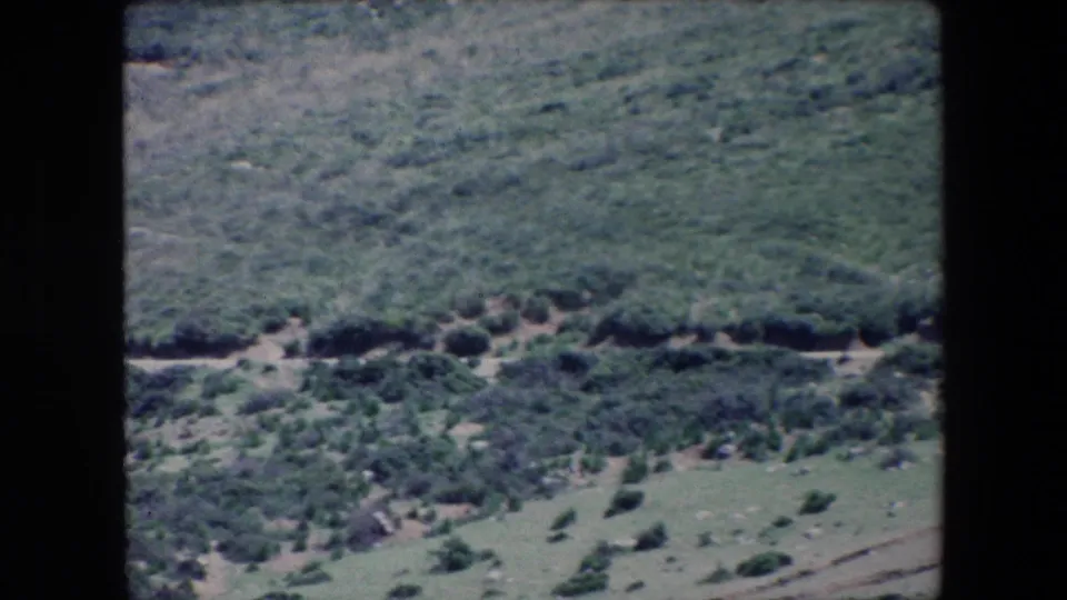 1971 Algeria 8mm frame of rugged, shrub-dotted hills where a narrow dirt road runs horizontally through the landscape.
