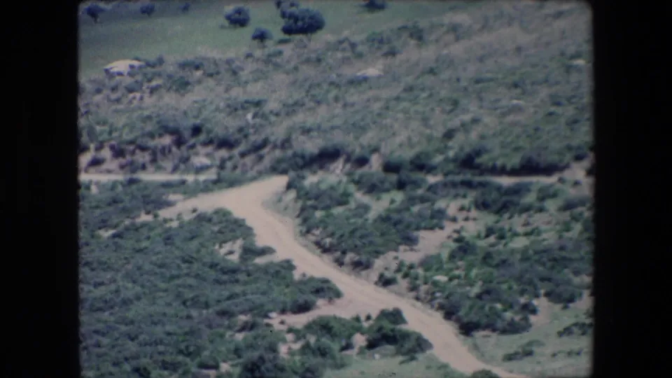 1971 Algeria 8mm frame with a winding pale dirt road bending through scrubland below a grassy, shrub-covered hillside.