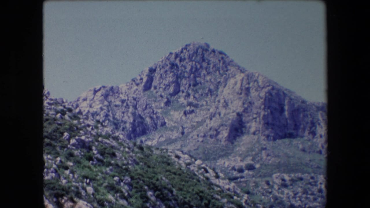 1971 Algeria 8mm frame showing a jagged rocky mountain above green scrub and a valley slope, with visible rounded film-frame edges.