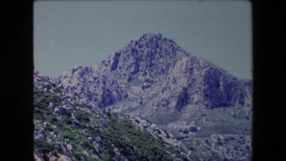 1971 Algeria 8mm frame showing a jagged rocky mountain above green scrub and a valley slope, with visible rounded film-frame edges.