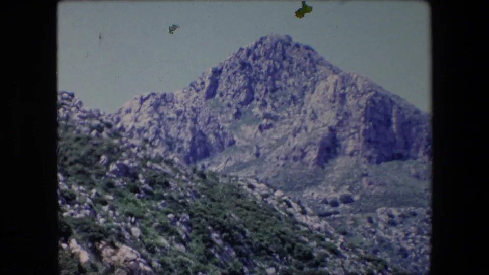 1971 Algeria 8mm frame with a rugged mountain peak, rocky hillside, and dense green shrubs viewed beneath a clear pale sky.