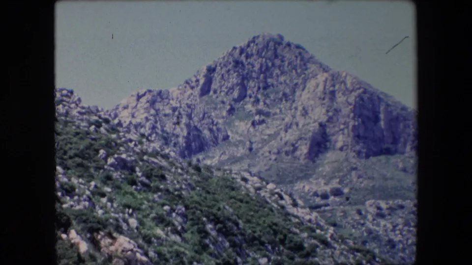 1971 Algeria 8mm frame showing the mountain face from a slightly lower composition, with scrub, boulders, and valley terrain in the foreground.