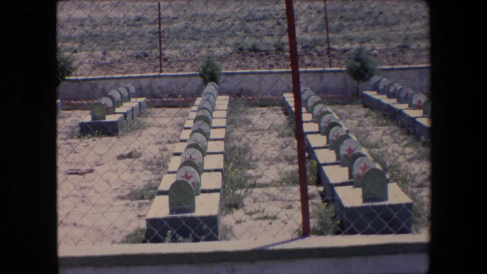 1971 Algeria 8mm frame showing rows of uniform grave markers with red stars behind chain-link fencing in a dry cemetery area.