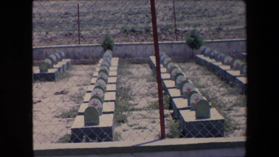 1971 Algeria 8mm frame showing a military-style cemetery with aligned grave markers, red star symbols, dry soil, and fence mesh in the foreground.