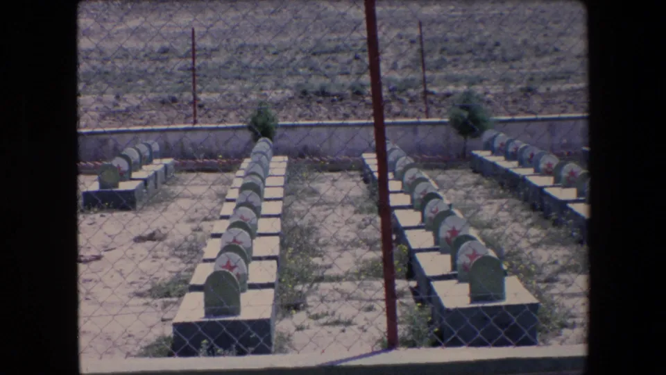 1971 Algeria 8mm frame showing repeated rows of red-star grave markers behind chain-link fencing with dry open ground beyond.