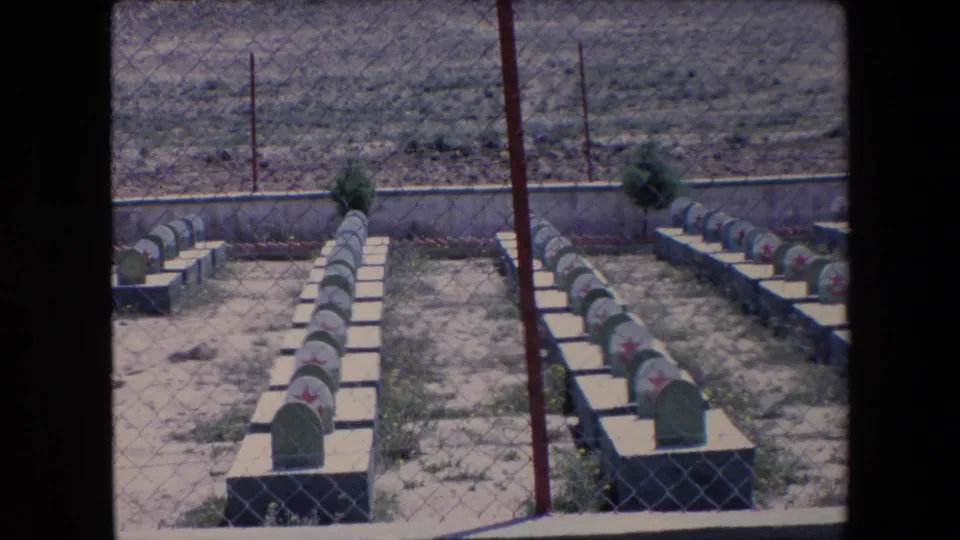 1971 Algeria 8mm frame showing rows of green grave markers with red star emblems behind a chain-link fence in an arid cemetery.