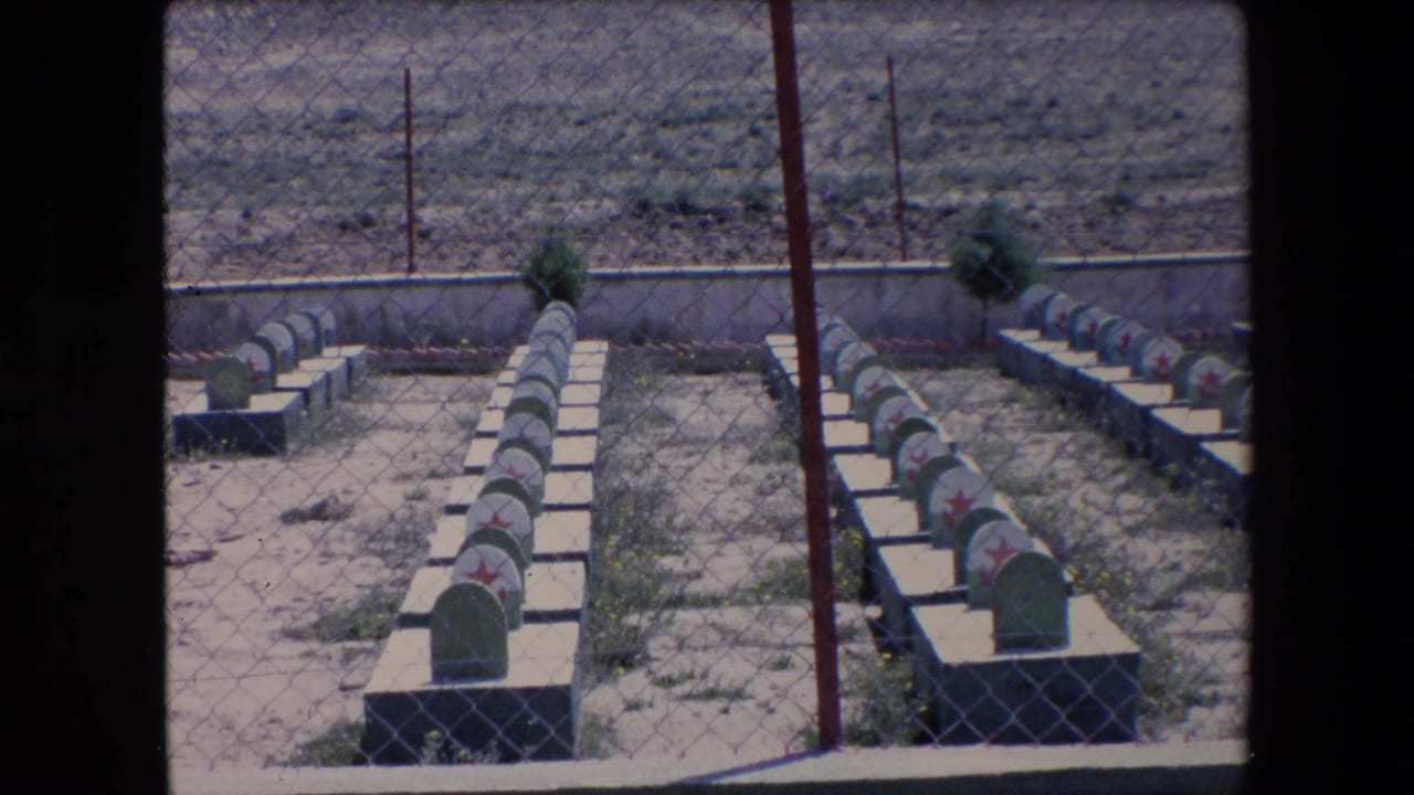 1971 Algeria 8mm frame of uniform green headstones with red star markings arranged in rows behind chain-link fencing and a low cemetery wall.