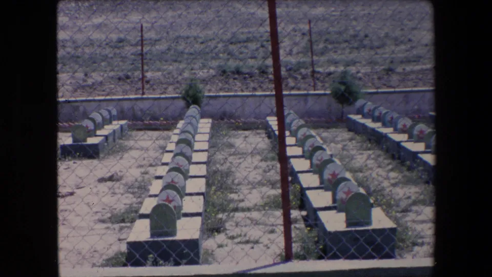 1971 Algeria 8mm frame of uniform green headstones with red star markings arranged in rows behind chain-link fencing and a low cemetery wall.