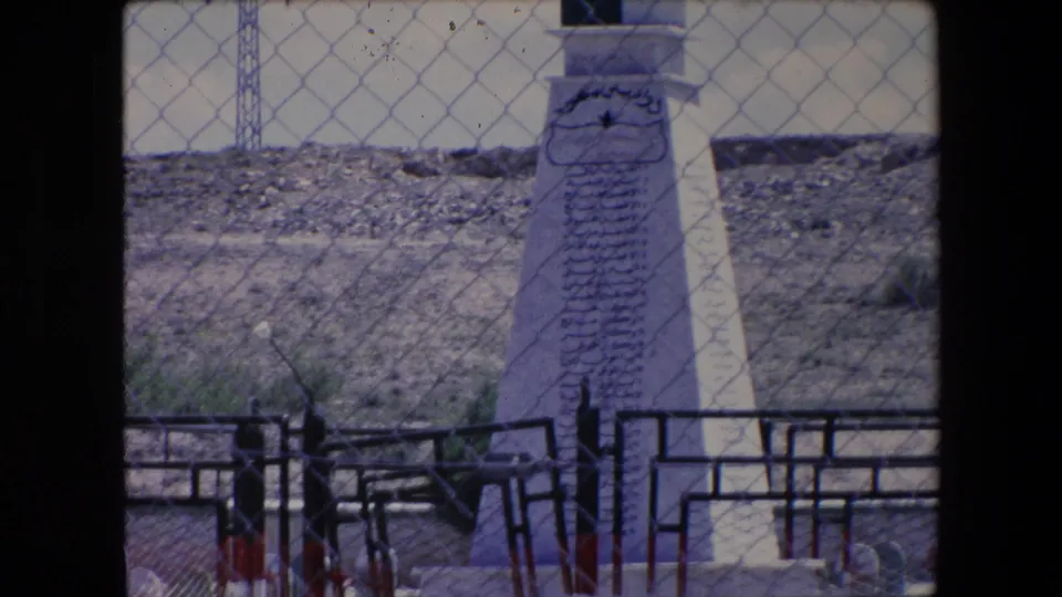 1971 Algeria 8mm frame showing a tall white memorial with Arabic inscriptions below fenced rows of green cemetery headstones.