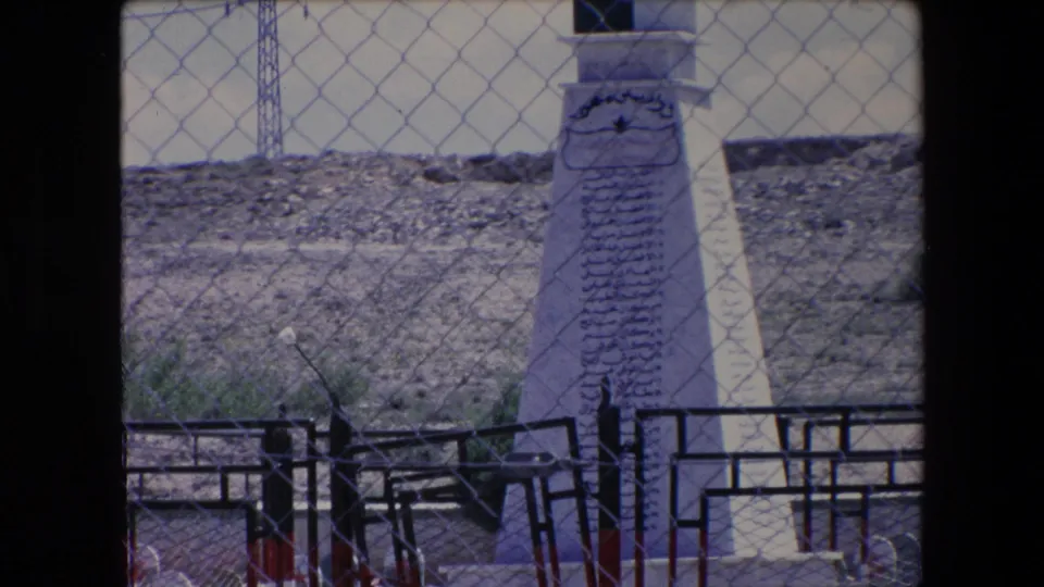 1971 Algeria 8mm frame with a white Arabic-inscribed memorial in the foreground and orderly green headstones behind a chain-link fence.