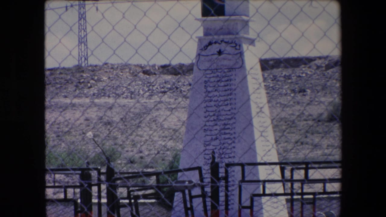 1971 Algeria 8mm frame showing a white stone monument with Arabic inscriptions, partly obscured by a chain-link fence and foreground railings in a rocky outdoor setting.