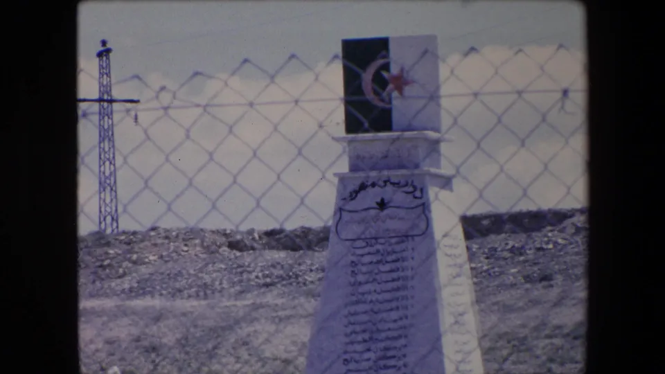 1971 Algeria 8mm frame showing the upper portion of a white monument with Arabic text and a crescent-and-star plaque, with chain-link fencing and a utility tower behind it.