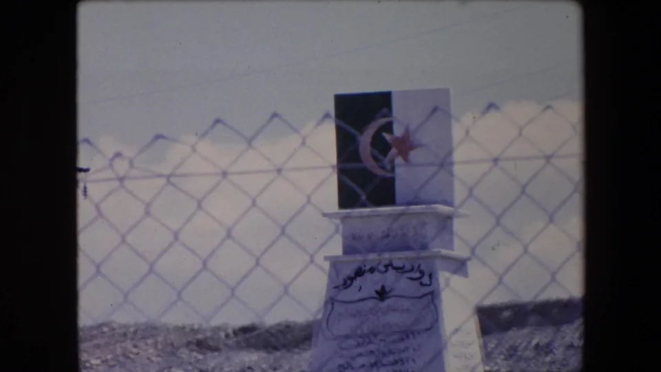 1971 Algeria 8mm frame focusing on the upper monument with a crescent-and-star emblem and Arabic inscription, seen through a soft chain-link fence pattern.