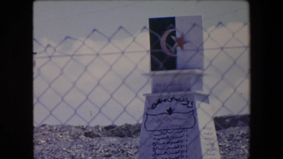 1971 Algeria 8mm frame showing the monument from a slightly lower angle, with Arabic inscriptions, a crescent-and-star plaque, chain-link fencing, rocky ground, and pale sky.