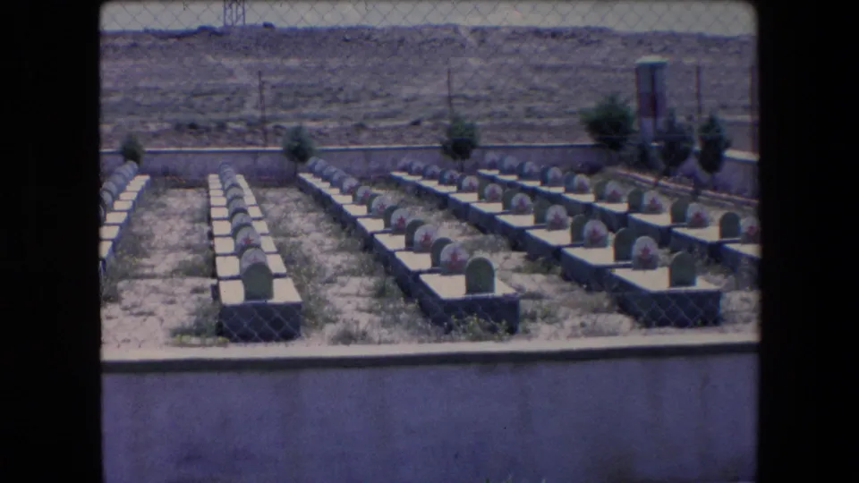 1971 Algeria home-movie frame showing rows of white grave bases with green headstones and red star markings viewed through a chain-link fence in an arid landscape.