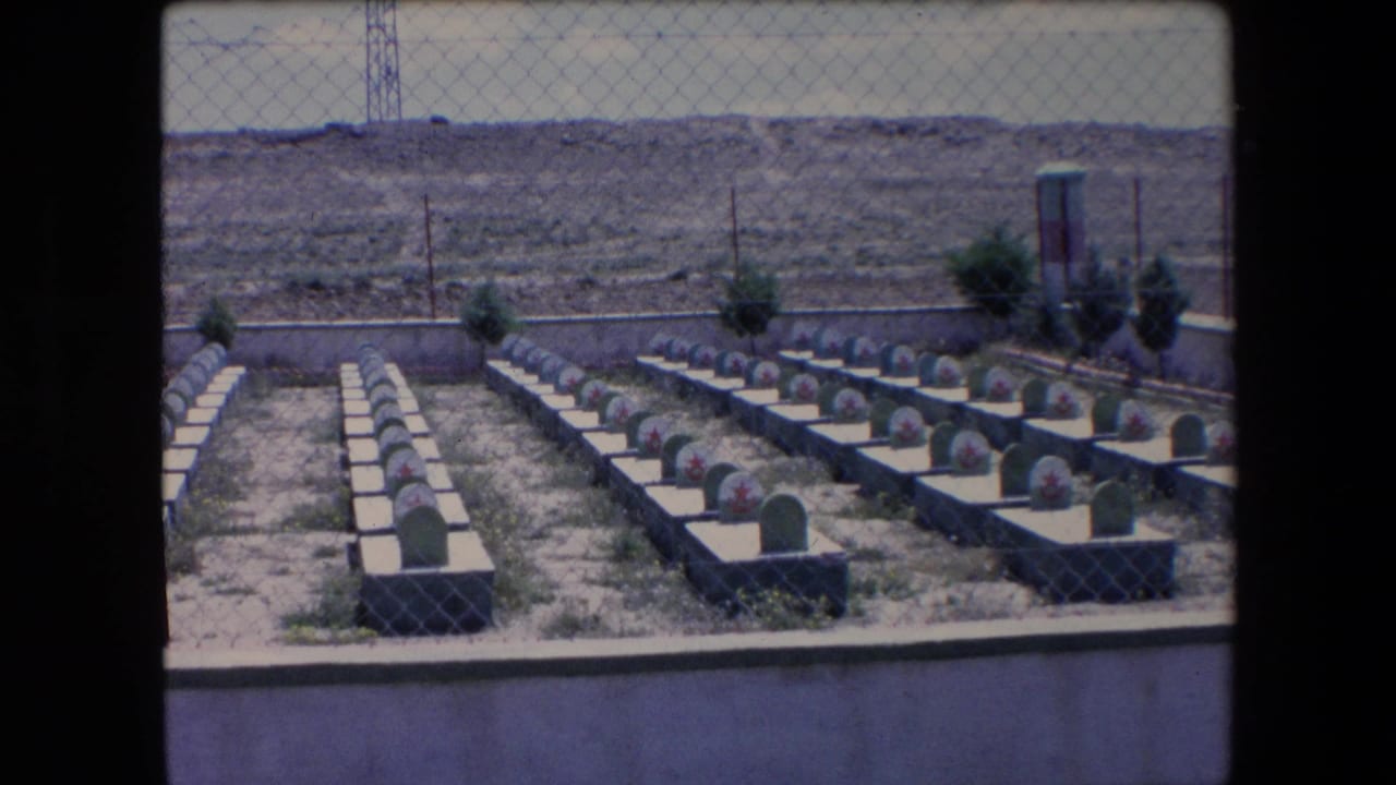 Uniform grave rows and a small monument or sign structure beside shrubs in a dry cemetery.