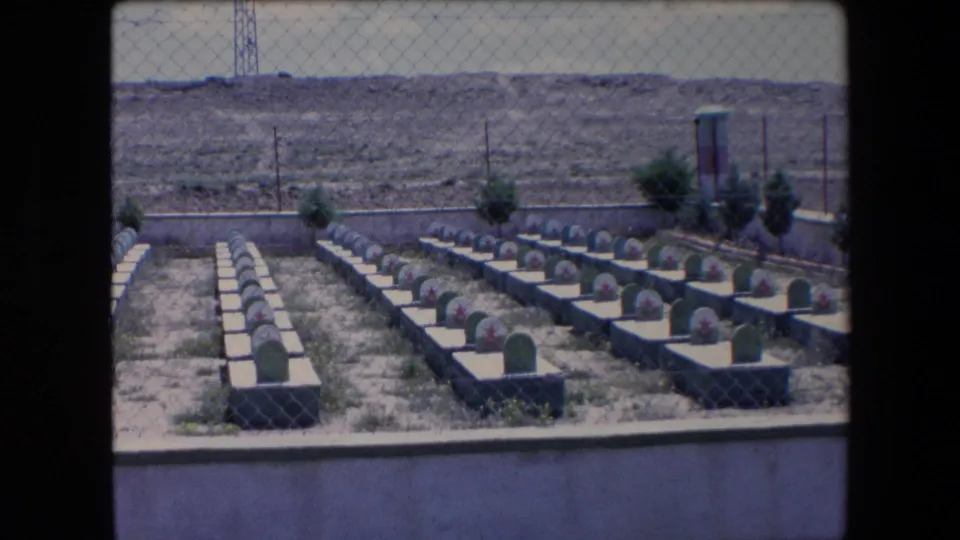 1971 Algeria 8mm still showing a fenced cemetery with orderly grave rows, rounded green headstones, red star markings, and sparse vegetation against a barren hillside.