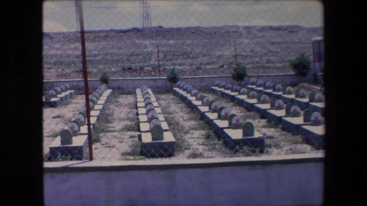 1971 Algeria 8mm view across a fenced cemetery, with repeated grave markers, red star emblems, a low perimeter wall, and barren terrain in the background.