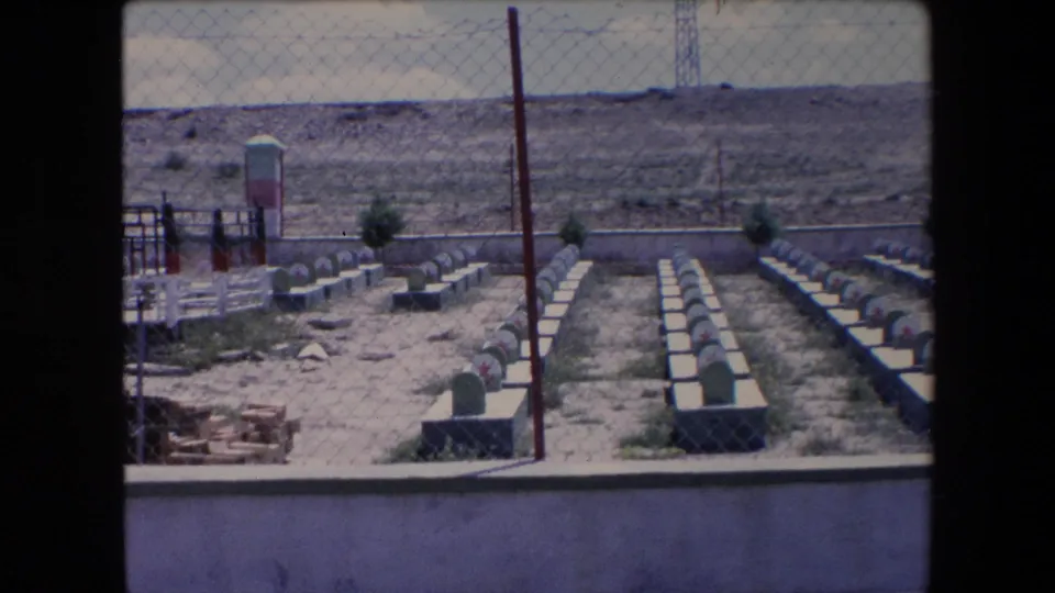 1971 Algeria 8mm frame showing the cemetery from behind a chain-link fence, with aligned headstones, dry soil, a vertical pole, and a small gated or monument area at left.