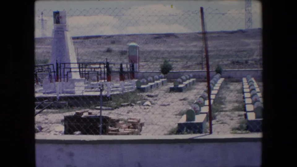 1971 Algeria 8mm frame showing a white obelisk-like cemetery monument, fenced grave rows with red-star markers, dry terrain, and low hills beyond.