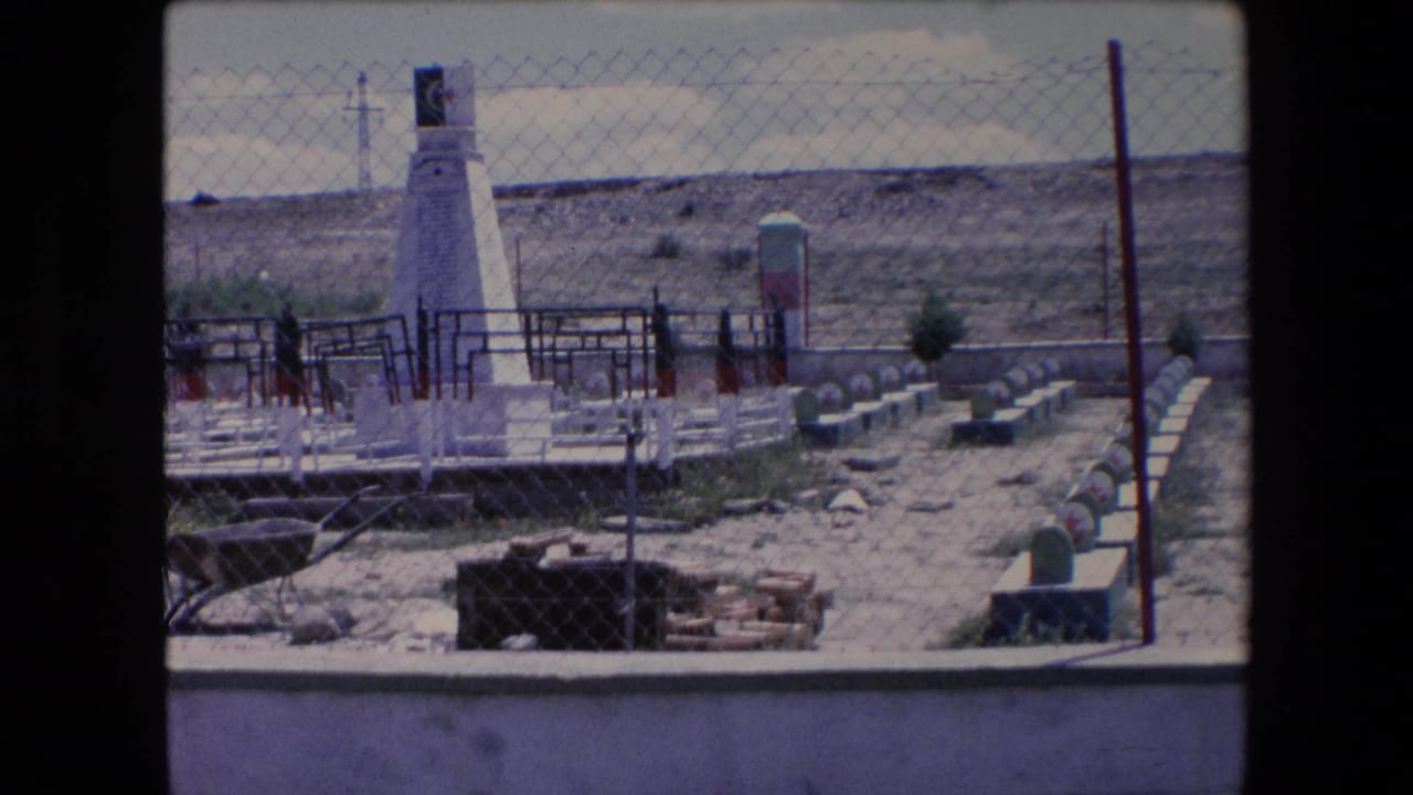 1971 Algeria 8mm frame showing a fenced memorial or cemetery-like enclosure with a white obelisk, green-topped markers, sparse vegetation, and an arid hillside beyond.