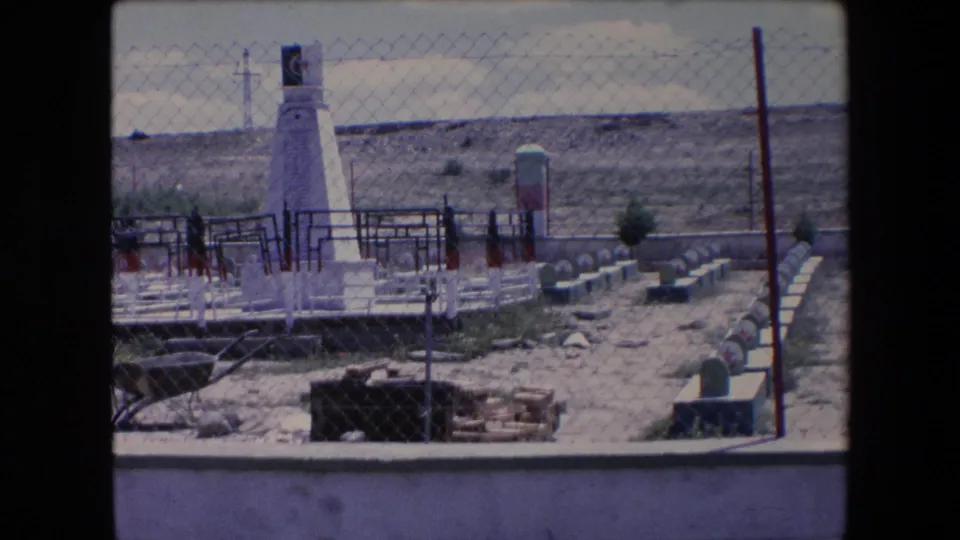 1971 Algeria 8mm frame showing a fenced memorial or cemetery-like enclosure with a white obelisk, green-topped markers, sparse vegetation, and an arid hillside beyond.