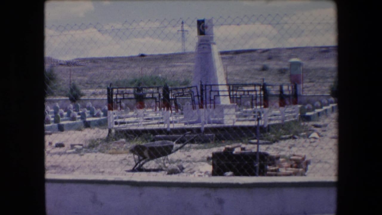 White obelisk behind chain-link fencing with grave markers, desert vegetation, and maintenance materials nearby.