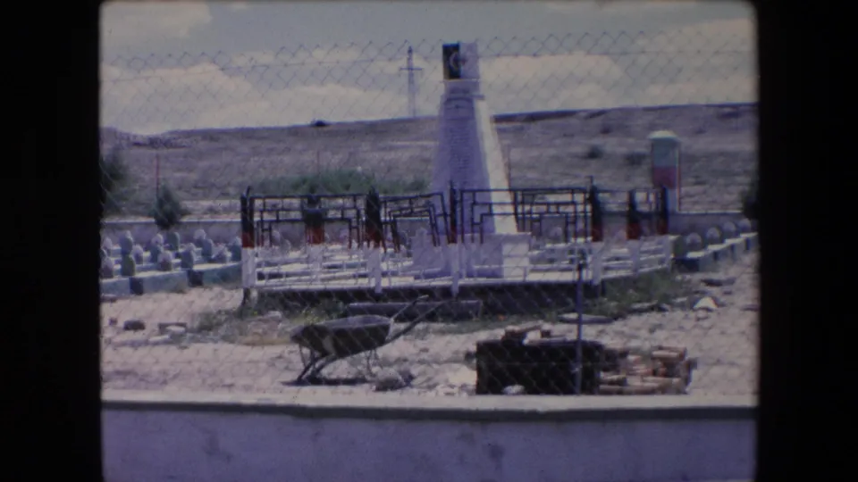 1971 Algeria 8mm view of a white obelisk and rows of small grave markers behind chain-link fencing, with dry terrain and a distant power line in the background.