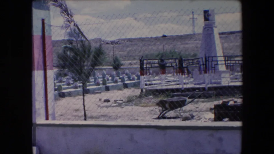 1971 Algeria 8mm frame looking through a chain-link fence toward a memorial enclosure with green-topped grave markers, a white obelisk, a small tree, and a wheelbarrow in the foreground.